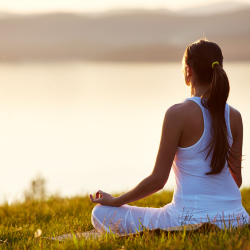 Woman sitting in meditative posture on grass in front of a lake.